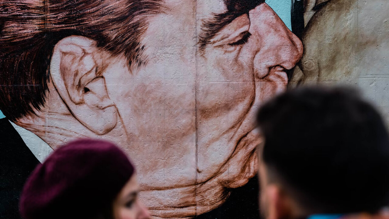 Monumentales Wandgem&auml;lde eines gek&uuml;ssten Gesichts, das die Berliner Mauer und eine historische politische Begegnung symbolisiert, mit verschwommenen Betrachtern im Vordergrund.