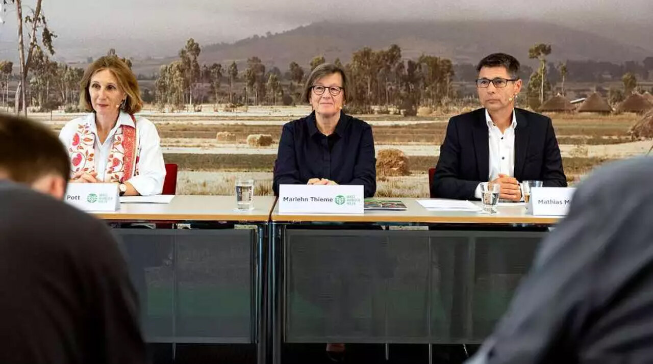 Drei Personen sitzen an einem Tisch vor einer Landschaftskulisse mit Palmen und Bergen, zwei Frauen und ein Mann in formeller Kleidung w&auml;hrend einer Konferenz oder Pressekonferenz.