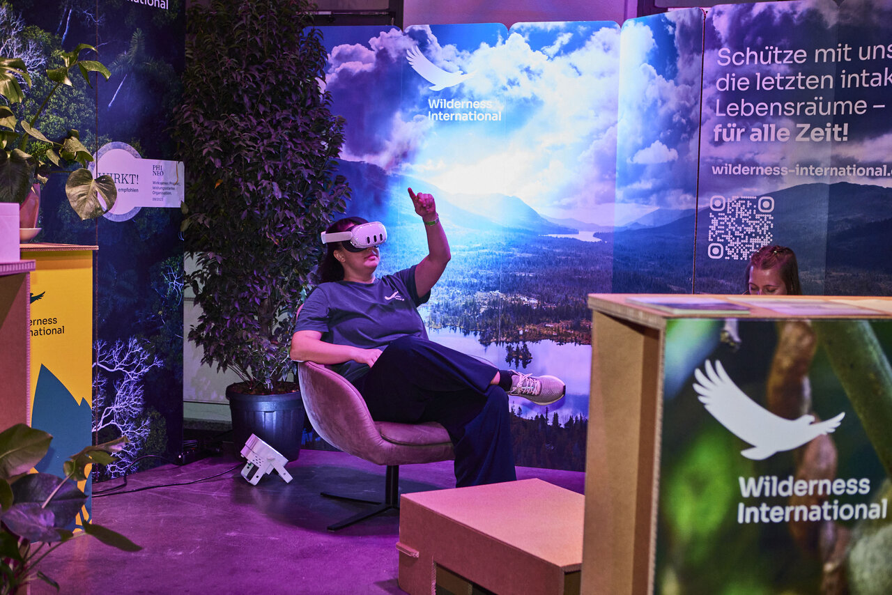 Eine Person sitzt in einem VR-Headset und interagiert mit einer virtuellen Umgebung auf einem Stand von Wilderness International, umgeben von einer atmosph&auml;rischen Kulisse mit Wolken und Naturmotiven.