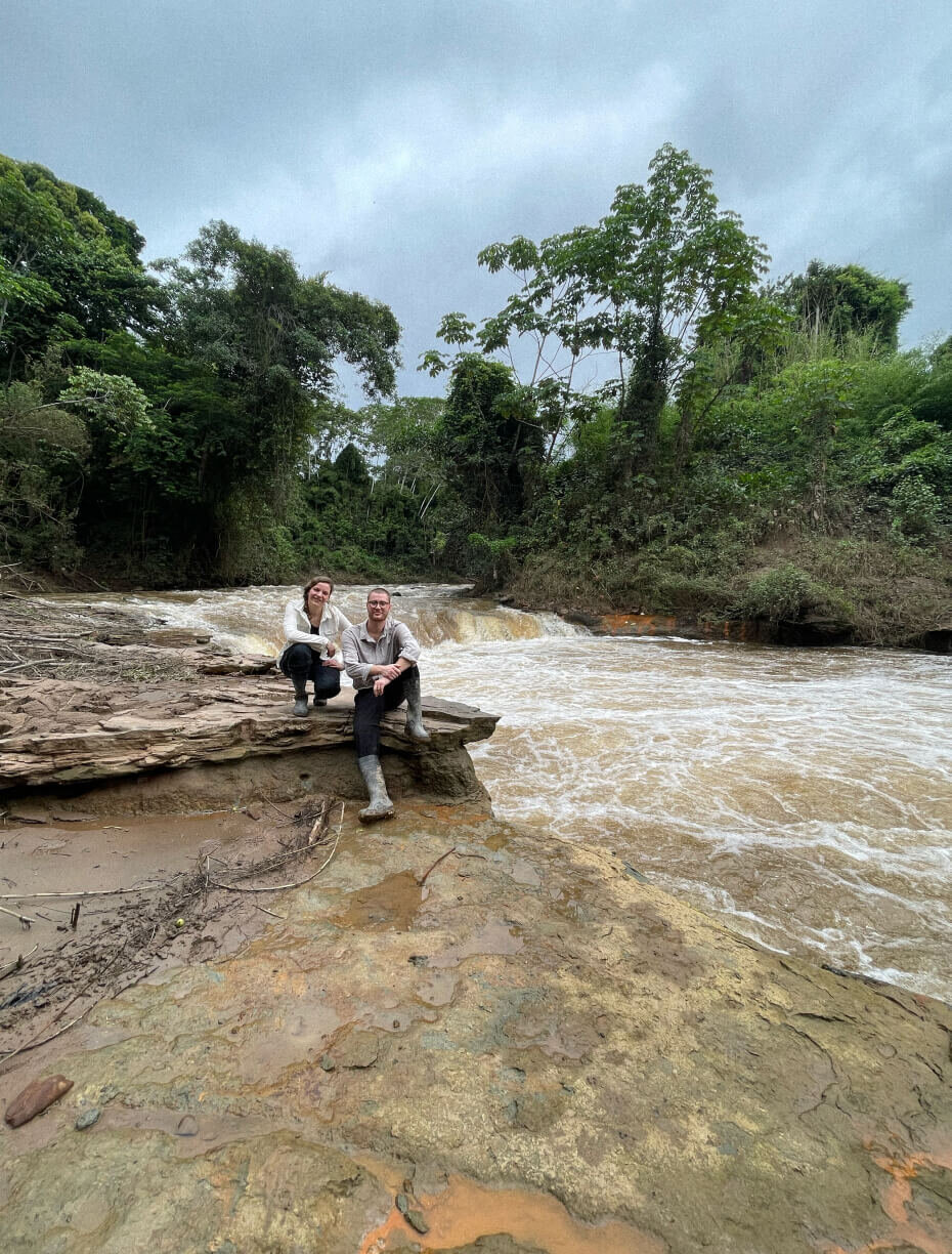 Zwei Personen sitzen auf einem Felsen an einem wilden, braunen Fluss, umgeben von &uuml;ppigem gr&uuml;nem Dschungelwald. Die Landschaft ist wild und naturbelassen, mit felsigem Ufer und starker Str&ouml;mung.