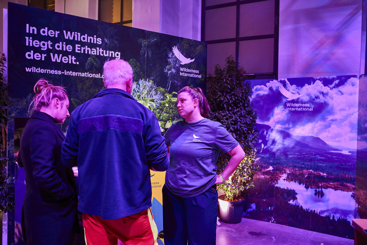 Drei Personen an einem Stand von Wilderness International mit einem Poster eines Waldsees, beleuchtet in violettem und blauem Licht, mit einem deutschsprachigen Banner &uuml;ber Naturerhaltung.