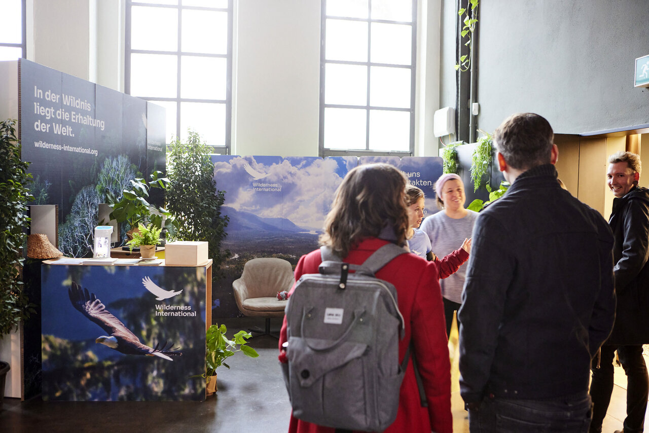 Drei Personen an einem Messestand mit Naturmotiven und Berglandschaft-Plakaten. Eine Person tr&auml;gt einen grauen Rucksack und eine rote Jacke, w&auml;hrend zwei weitere in einer Diskussion stehen. Der Stand ist mit Pflanzen und Informationstafeln dekoriert.