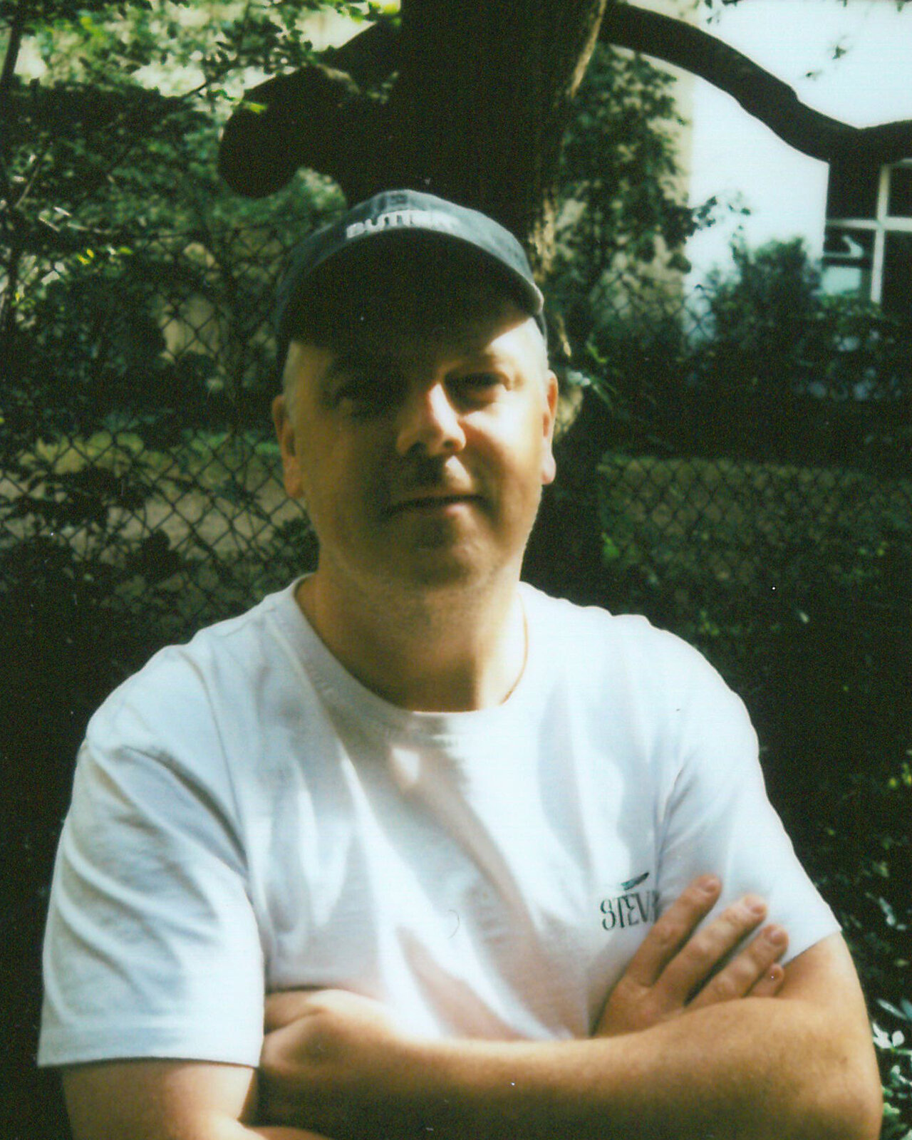 Ein Mann mit wei&szlig;em T-Shirt und Kappe steht vor einem Baum und einem Gartenzaun, die Arme verschr&auml;nkt, mit ruhigem Gesichtsausdruck.