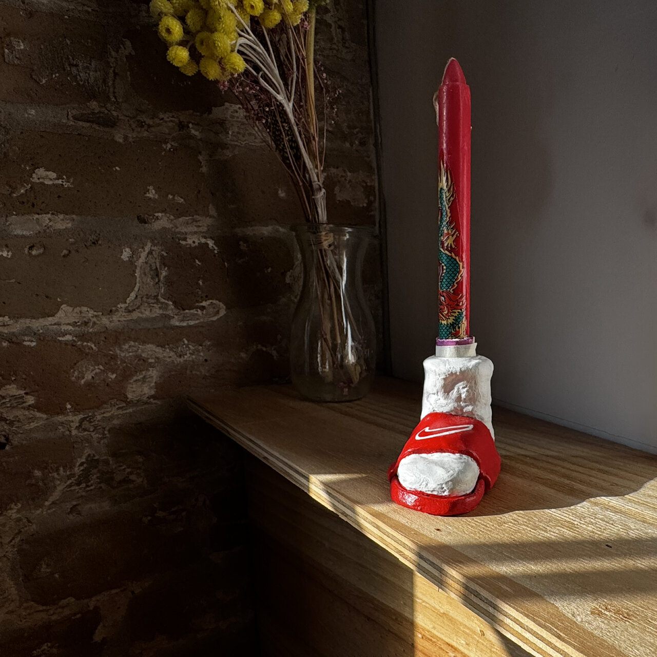 Rote Kerze auf wei&szlig;em Kerzenhalter vor einer Backsteinwand, daneben eine Vase mit gelben Blumen. Warmes Sonnenlicht f&auml;llt auf ein h&ouml;lzernes Regal und schafft eine stimmungsvolle, atmosph&auml;rische Beleuchtung.