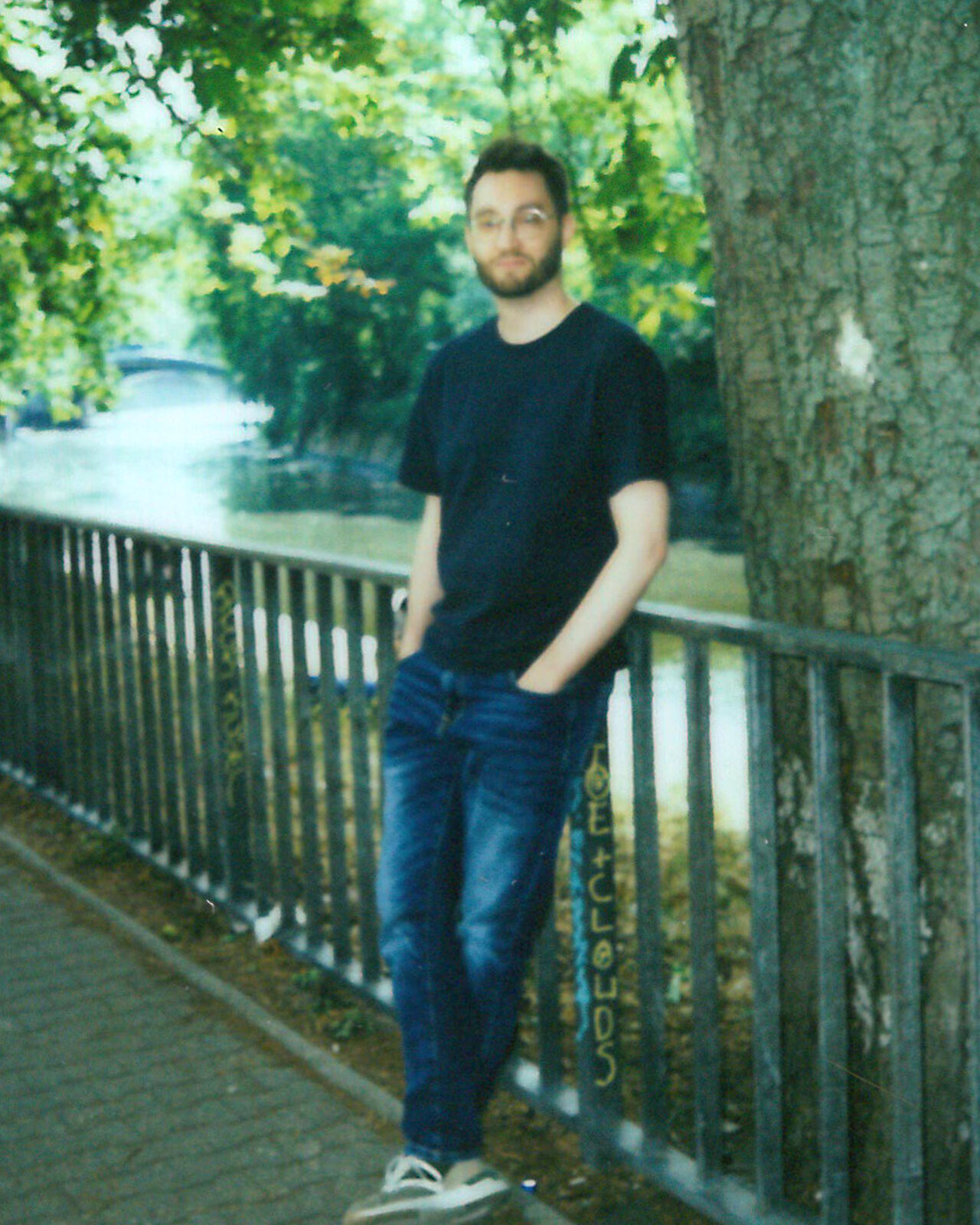Person in dunklem T-Shirt und blauen Jeans, die an einem Gel&auml;nder an einem Flussufer mit gr&uuml;nen B&auml;umen und verschwommenem Hintergrund lehnt. Er tr&auml;gt Turnschuhe und blickt leicht l&auml;chelnd in die Kamera.
