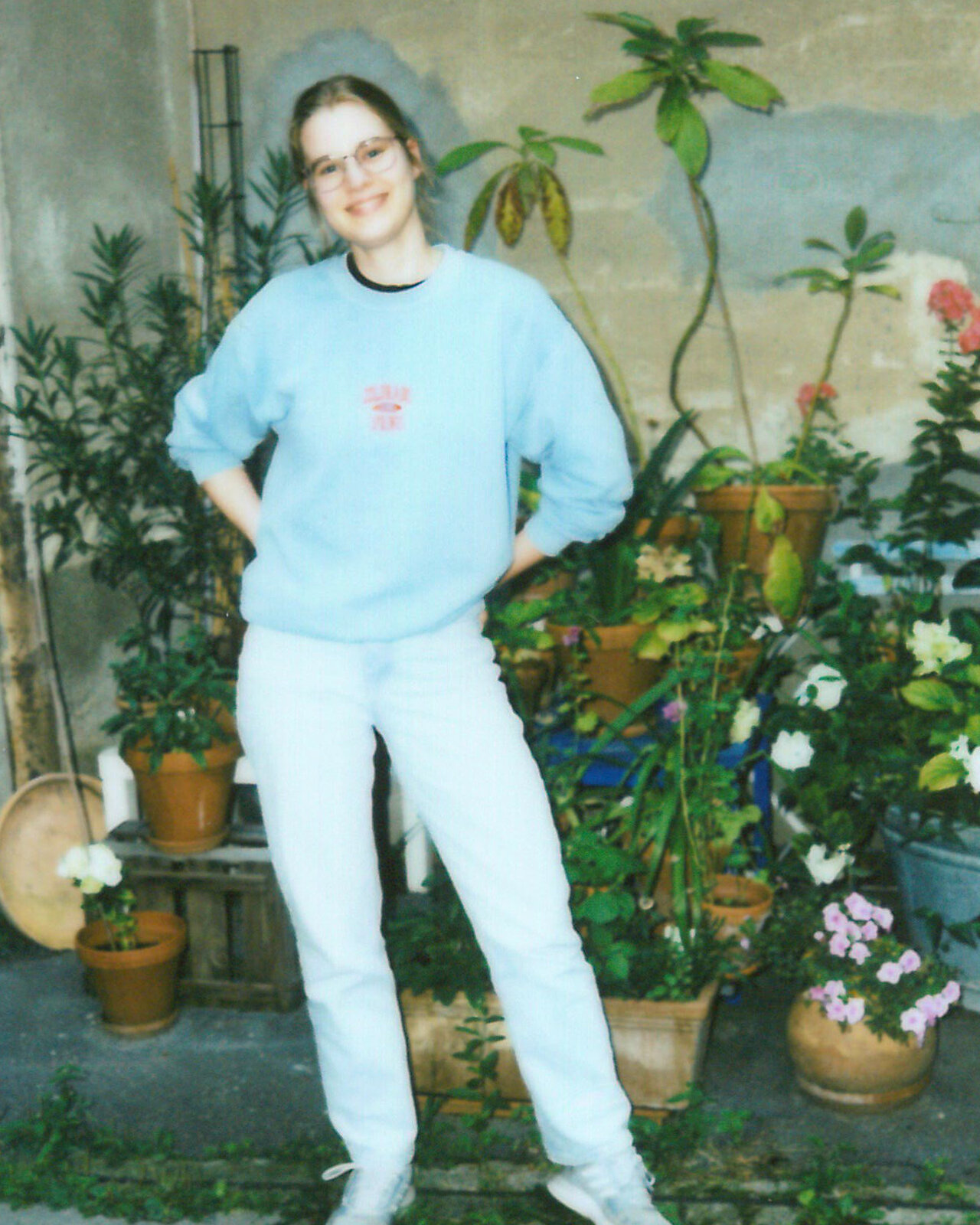 Eine Mitarbeiterin von Studio Good l&auml;chelt in die Kamera und tr&auml;gt einen hellblauen Pullover. Sie steht vor einer gr&uuml;nen Kulisse mit Bl&auml;ttern und Zweigen, was einen nat&uuml;rlichen, entspannten Hintergrund bildet.