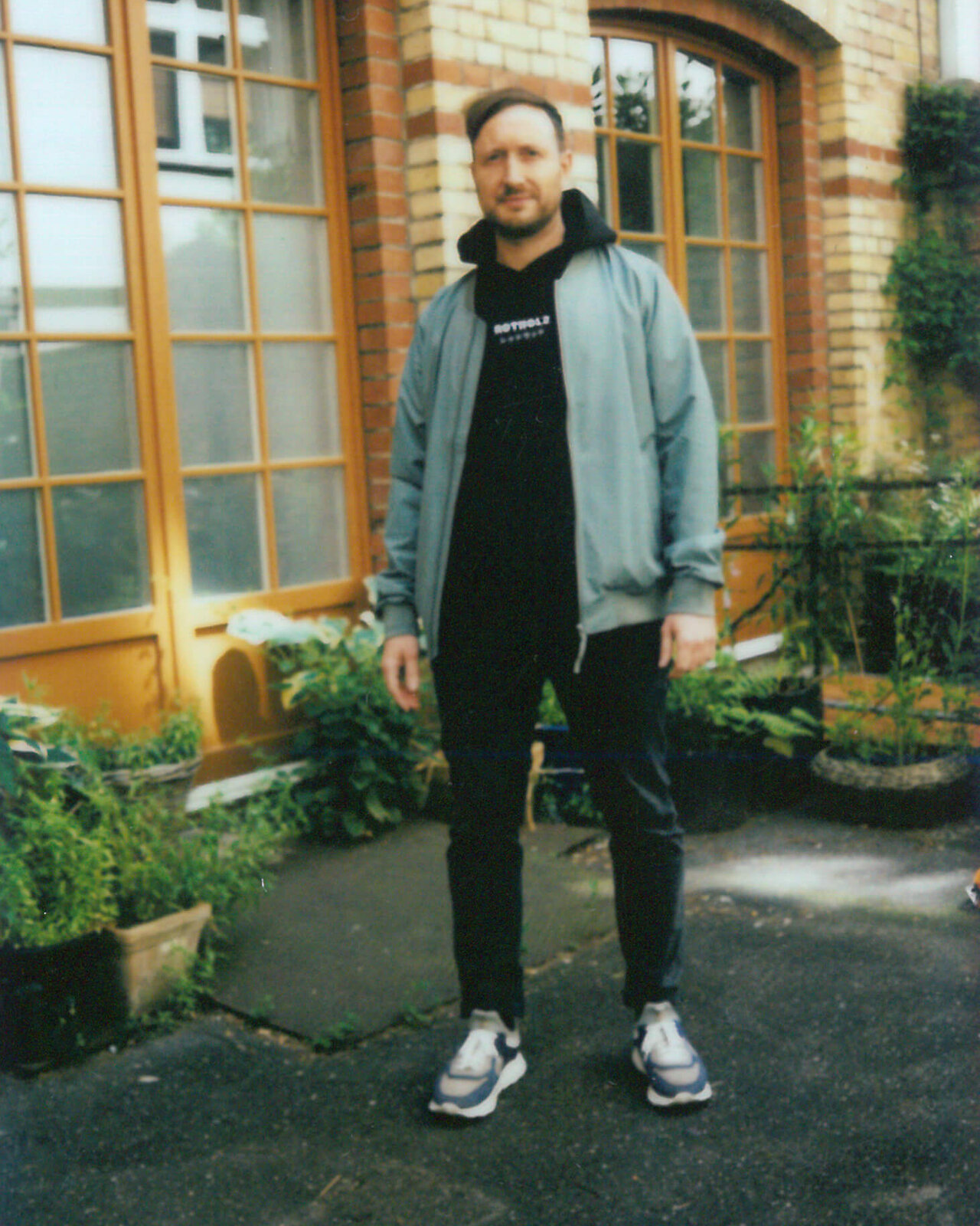 Ein Mitarbeiter von Studio Good steht vor einem Backsteingeb&auml;ude mit Holzfenstern, gekleidet in eine graue Jacke, schwarzer Hoodie und Sneaker. Er posiert vor einer Pflanzenwand und scheint sich wohl zu f&uuml;hlen in der Umgebung des B&uuml;ros.