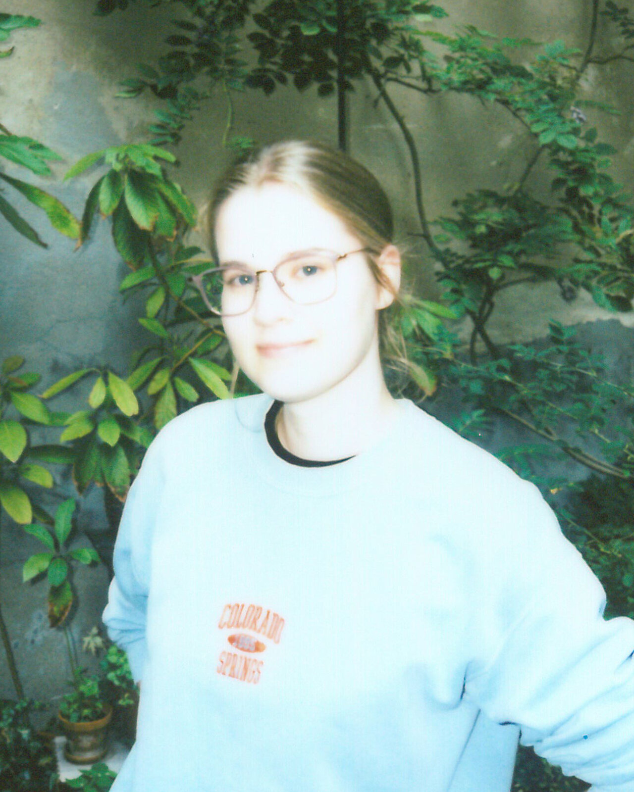 Eine Mitarbeiterin von Studio Good l&auml;chelt in die Kamera und tr&auml;gt einen hellblauen Pullover. Sie steht vor einer gr&uuml;nen Kulisse mit Bl&auml;ttern und Zweigen, was einen nat&uuml;rlichen, entspannten Hintergrund bildet.