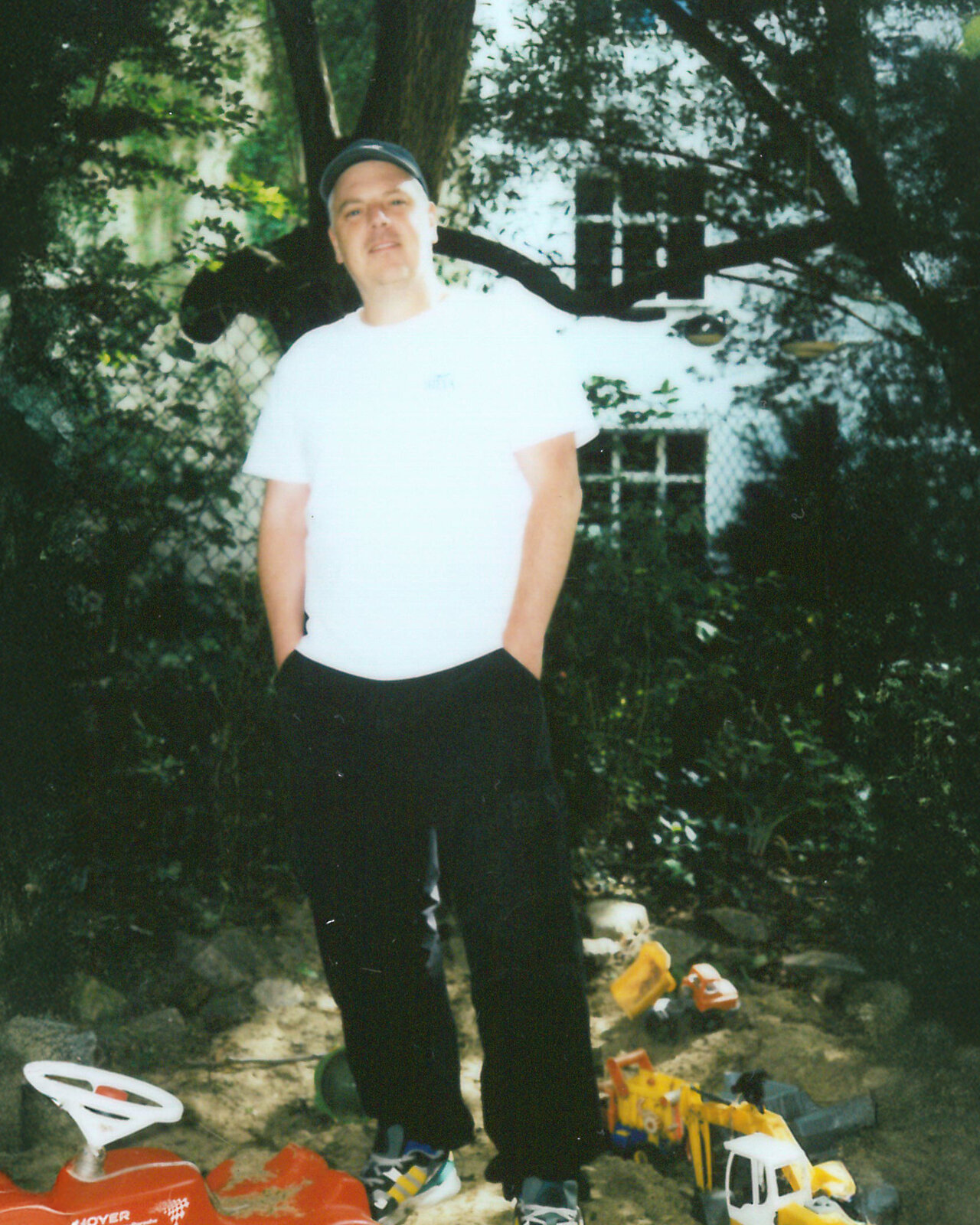 Ein Mann in wei&szlig;em T-Shirt und schwarzer Hose steht l&auml;ssig in einem gr&uuml;nen Garten, umgeben von Spielzeugautos und Kinderutensilien.