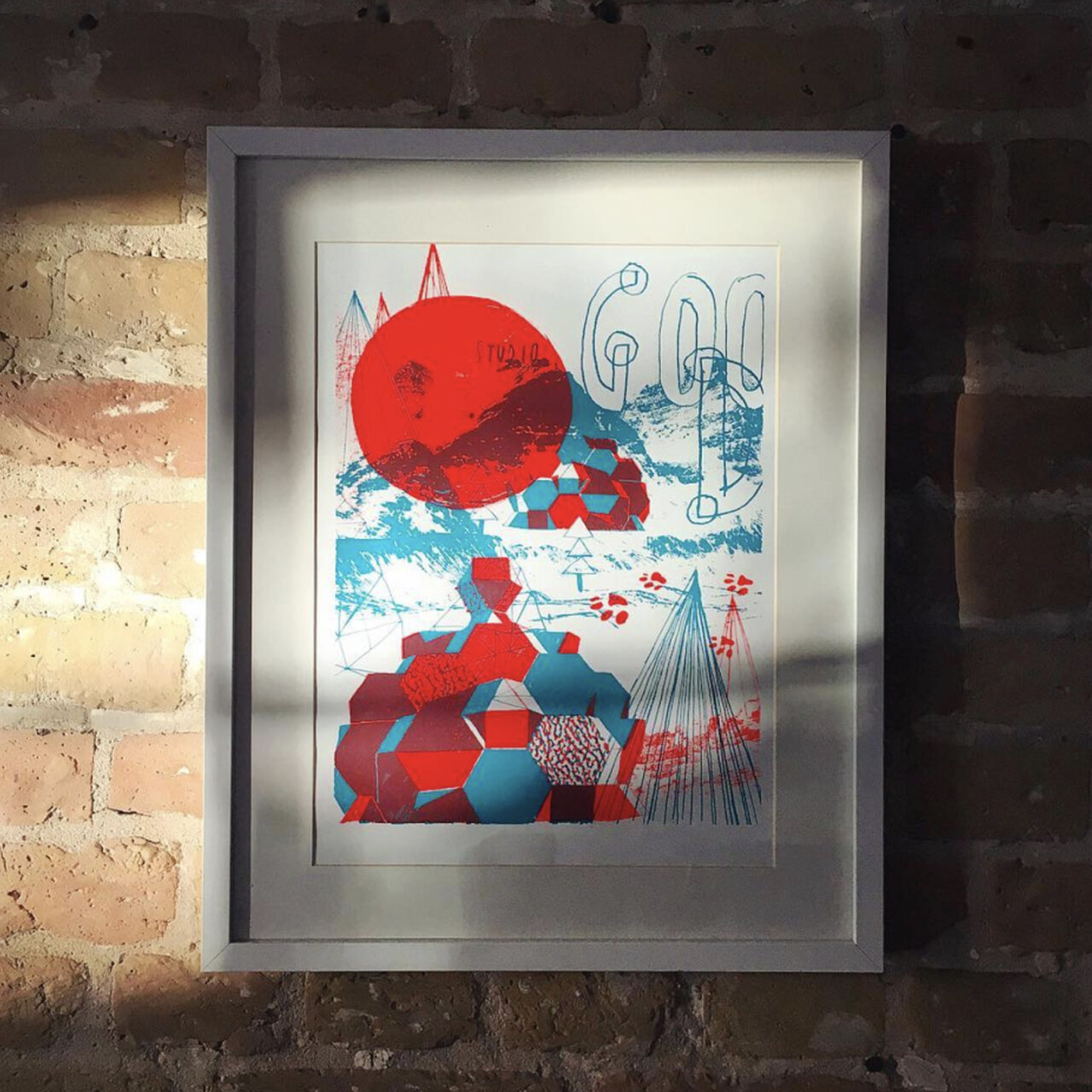 Abstrakter Kunstdruck in Rot und Blau mit geometrischen Formen wie Kugeln, Pyramiden und unregelm&auml;&szlig;igen Strukturen. Der Druck ist in einem grauen Rahmen vor einer Ziegelwand montiert, was einen interessanten Kontrast zwischen moderner Kunst und rustikaler Architektur schafft.
