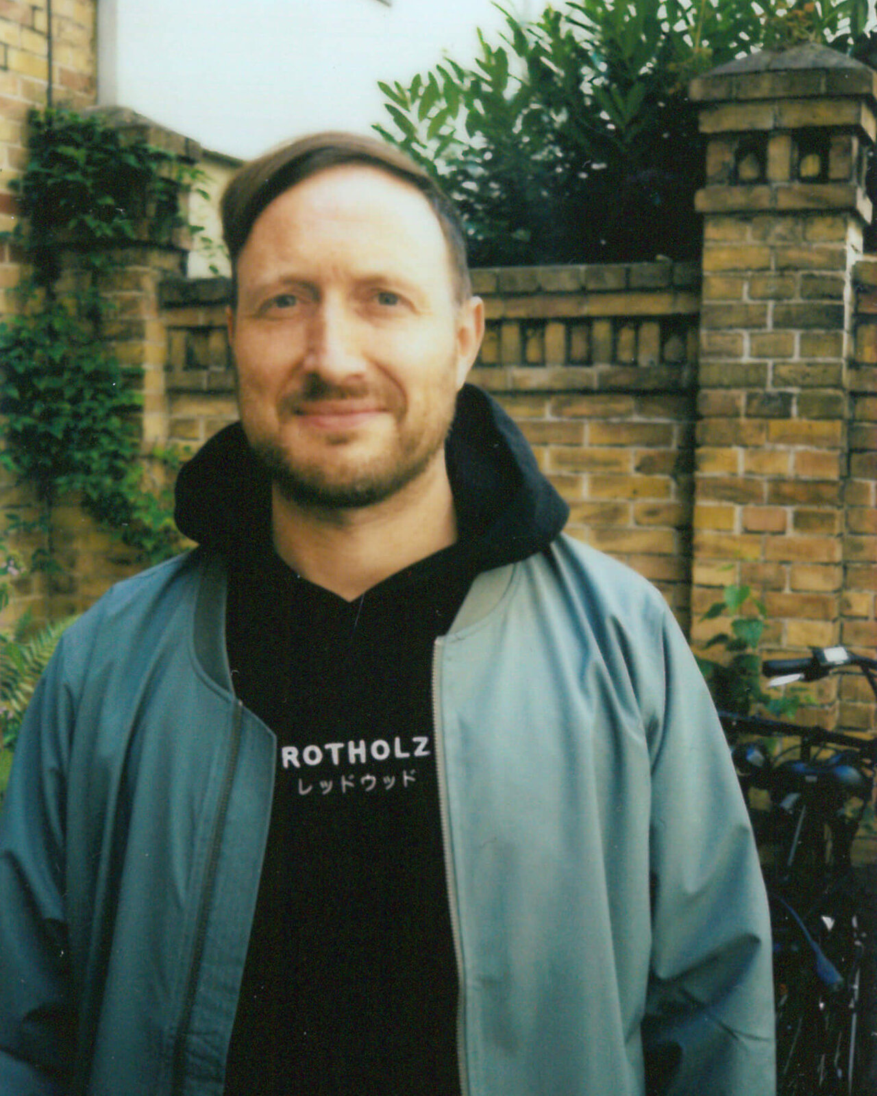 Mitarbeiter von Studio Good in Berliner Innenhof, gekleidet in schwarzem Rotholz-Hoodie und grauer Jacke, steht vor einer Backsteinmauer mit Efeu und Pflanzen im Hintergrund.