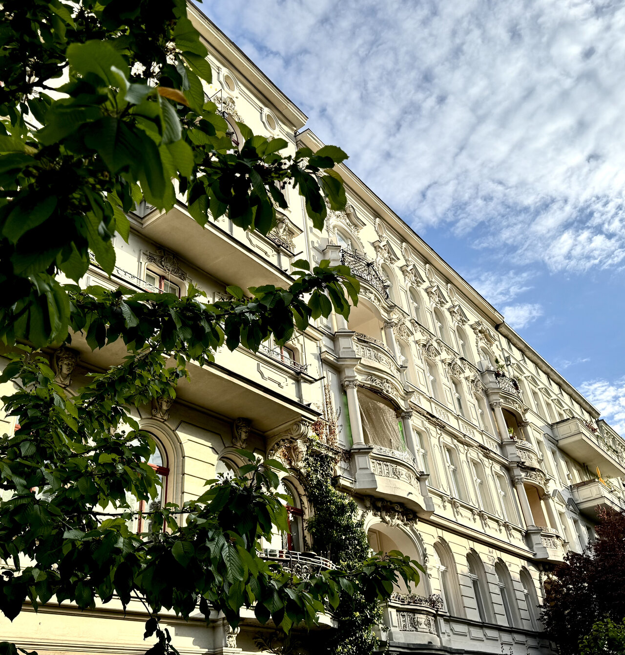 Historische Altbaufassade in Berlin mit Stuck und Verzierungen, wei&szlig;-gelb gestrichen, davor gr&uuml;ne Baumzweige vor blauem Wolkenhimmel. Klassische Architektur mit Erkerfenstern und Balkonen, typisch f&uuml;r das Berliner Stadtbild.