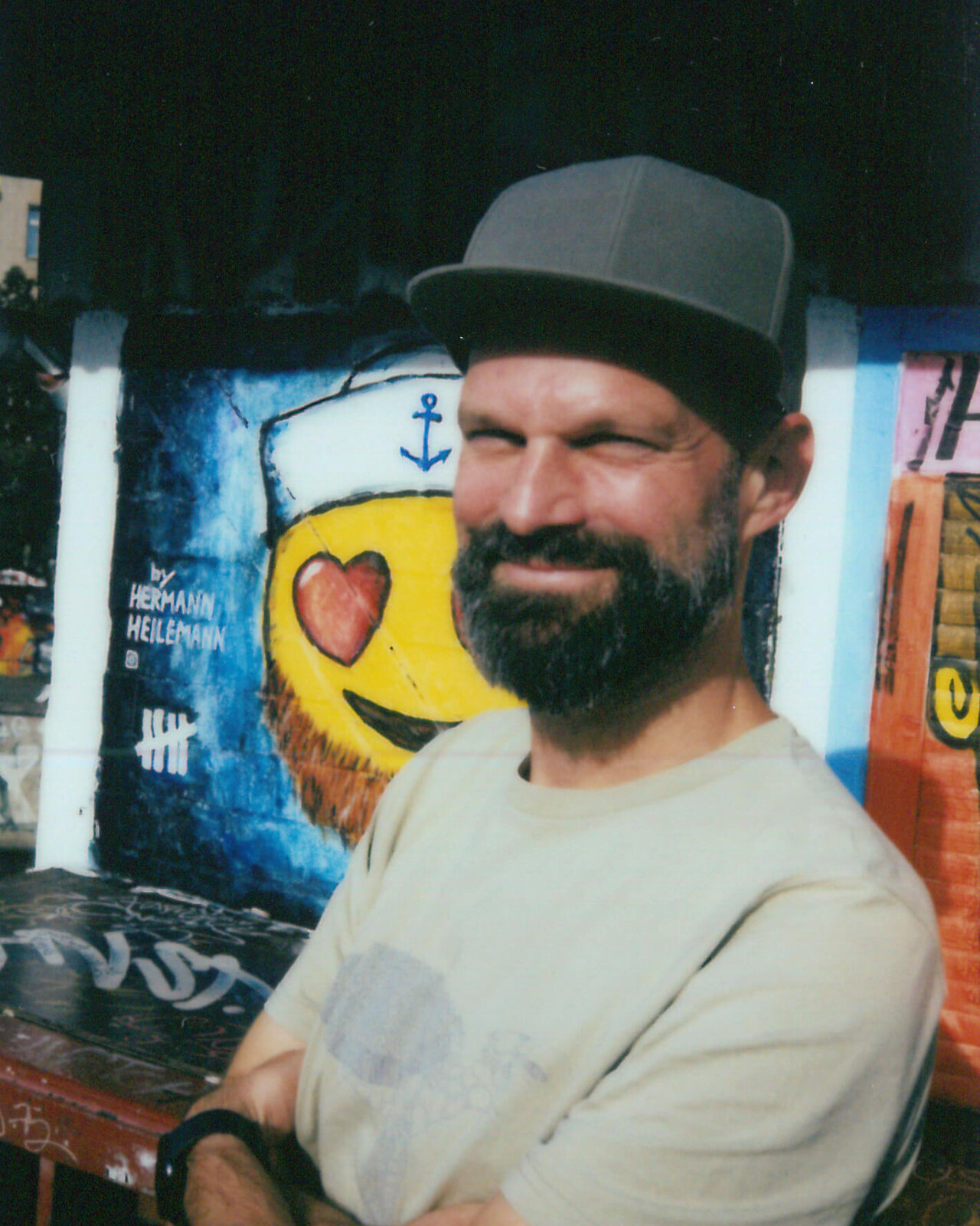 B&auml;rtiger Mann mit Basecap und wei&szlig;em T-Shirt, der vor einem bunten Graffiti-Wandhintergrund mit verschr&auml;nkten Armen l&auml;chelt. Er steht in einer entspannten Pose vor einer Wand mit Stra&szlig;enkunst, die ein Herz und einen Anker zeigt.