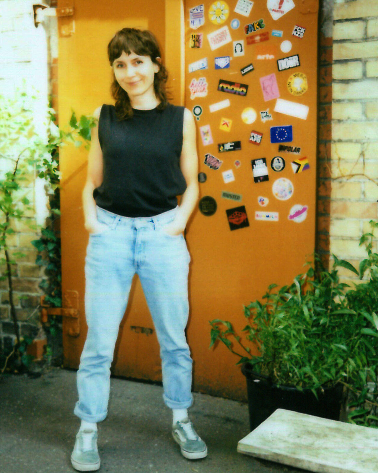 Eine Person in schwarzem Top und hellblauen Mom-Jeans steht vor einer orangefarbenen T&uuml;r mit bunten Stickern. Sie tr&auml;gt graue Sneaker und hat einen freundlichen Gesichtsausdruck, w&auml;hrend Gr&uuml;npflanzen und eine Steinbank den Hintergrund bilden.