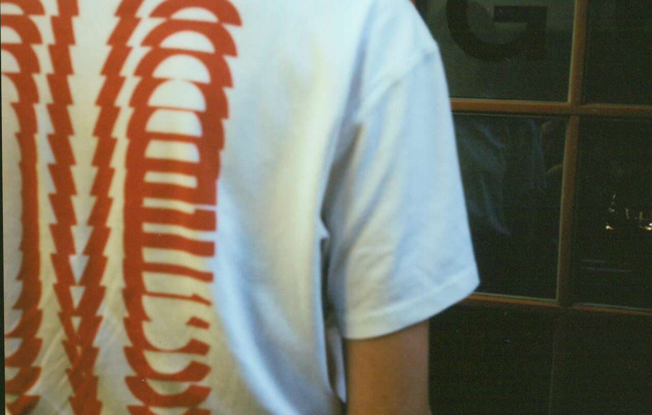 Wei&szlig;es T-Shirt mit orangefarbener, abstrakter Grafik an der Seite, die wellenf&ouml;rmige, sich verj&uuml;ngende Linien zeigt. Teilweise verschwommener Hintergrund mit dunkler Wand und Reflexion.