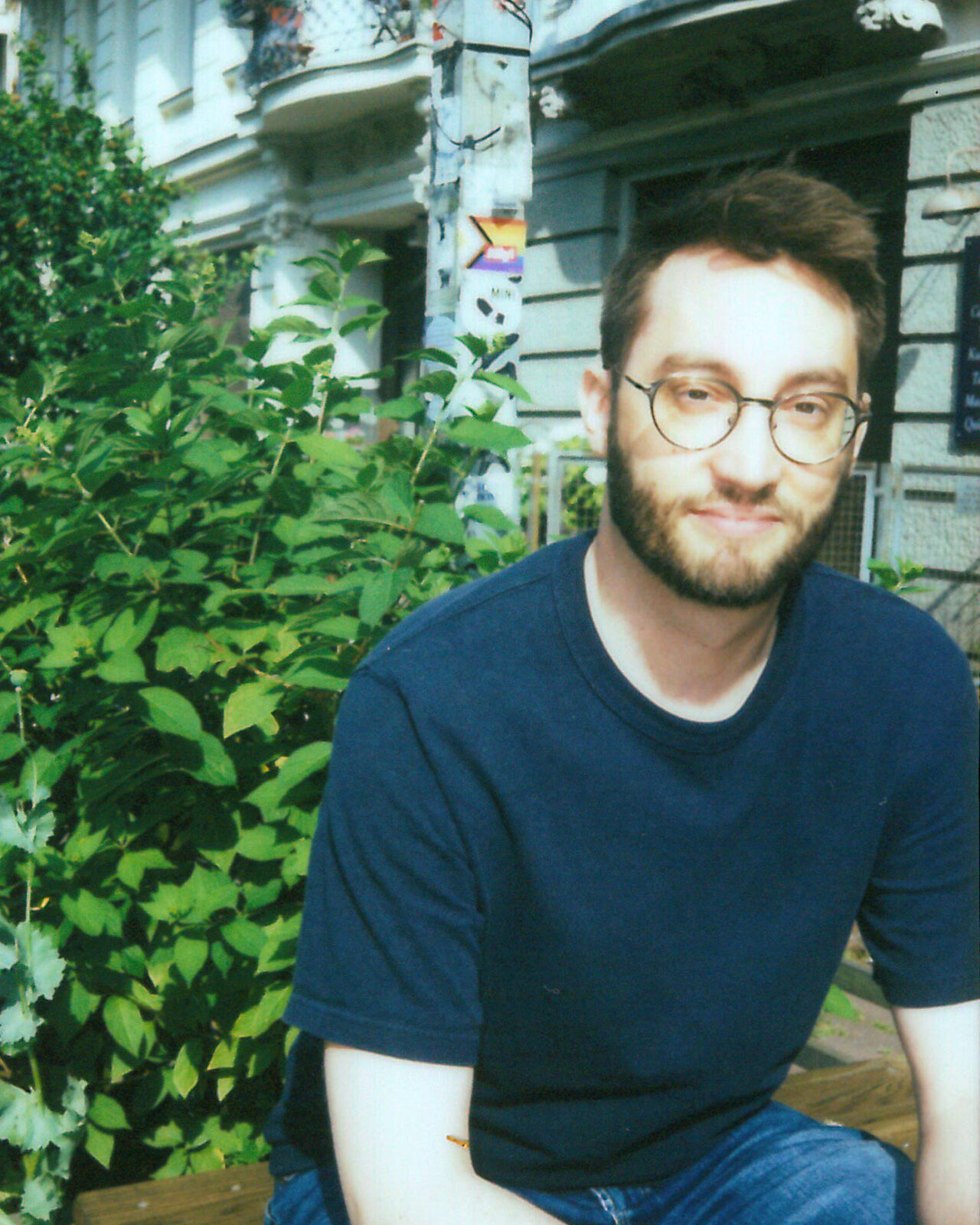 Junger Mann mit Brille und blauem T-Shirt sitzt auf einer Bank vor einem Geb&auml;ude mit Efeu und Blumen. Er tr&auml;gt einen Bart und l&auml;chelt leicht, w&auml;hrend er entspannt posiert. Die Umgebung wirkt gr&uuml;n und sommerlich mit einer historischen Architektur im Hintergrund.