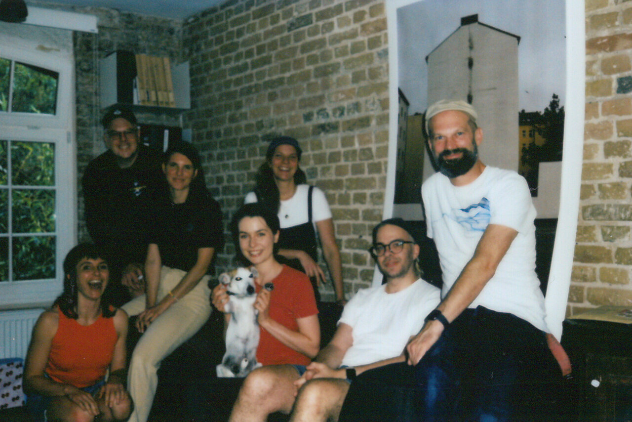 Sieben Personen in einem Ziegelsteinraum, sitzen und stehen zusammen, eine Person h&auml;lt einen Hund. Eine entspannte B&uuml;roatmosph&auml;re bei Studio Good, die Teamzusammenhalt und Kreativit&auml;t ausstrahlt.