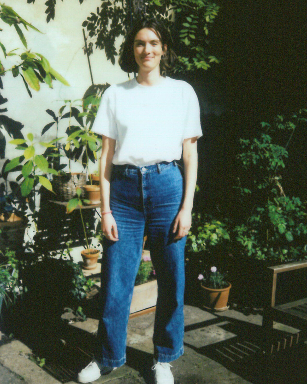 Eine Person in wei&szlig;em T-Shirt und blauen Jeans steht in einem Hinterhof mit Topfpflanzen und l&auml;chelt in die Kamera. Sie tr&auml;gt wei&szlig;e Turnschuhe und hat schulterlange Haare.