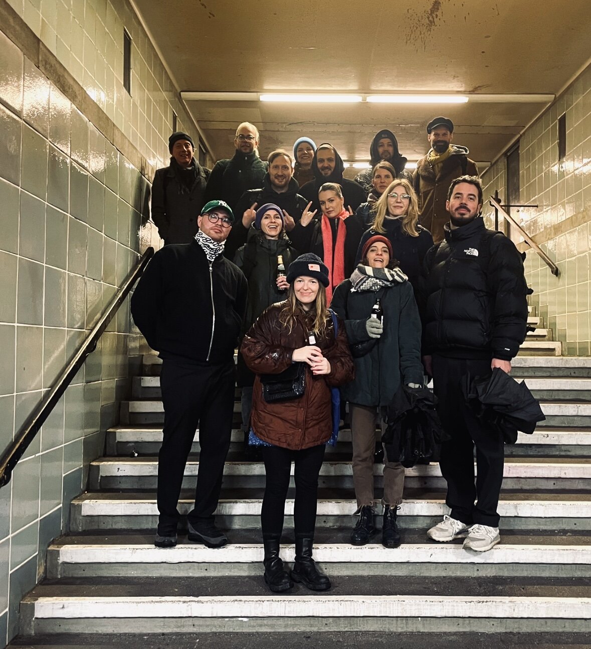 Eine Gruppe von etwa 15 Personen steht auf einer Treppe in einem Treppenhaus mit gefliesten W&auml;nden. Alle tragen Winterkleidung in dunklen Farben und stehen eng beieinander f&uuml;r ein Gruppenfoto.