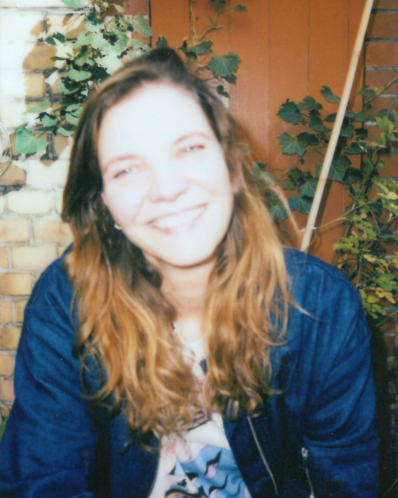 Junge Frau mit welligen, braunen Haaren l&auml;chelt in die Kamera. Sie tr&auml;gt eine blaue Jacke und ein gemustertes T-Shirt und sitzt vor einer Ziegelwand mit Efeu. Das Foto wirkt warm und pers&ouml;nlich, aufgenommen in entspannter Atmosph&auml;re.
