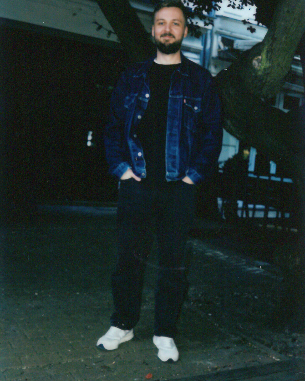 Ein Mann mit Bart tr&auml;gt eine blaue Jeansjacke und steht vor einem Baum und Geb&auml;uden. Er hat eine schwarze Hose und wei&szlig;e Turnschuhe und l&auml;chelt leicht in die Kamera.