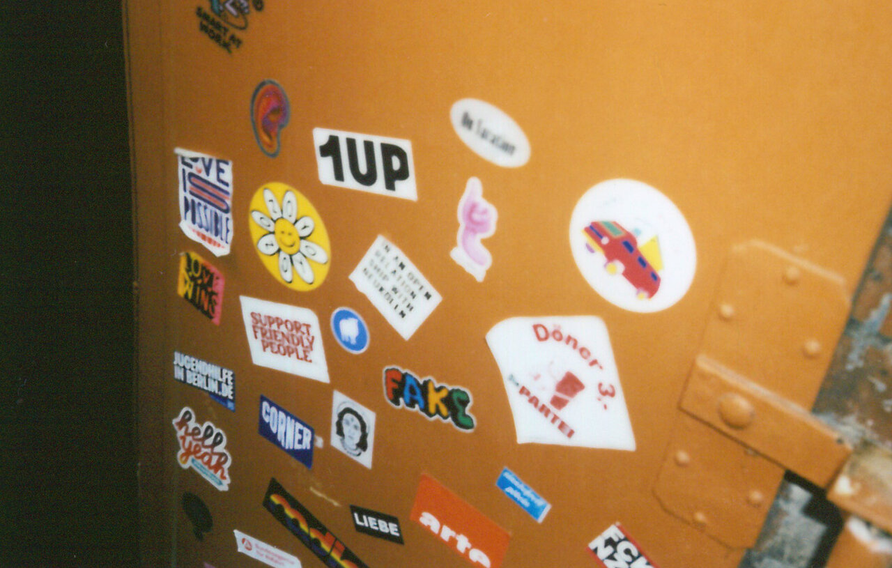 Gelbe Wand mit zahlreichen bunten Stickern und Aufklebern verschiedener Designs, darunter Logos, Grafiken, Schriftz&uuml;ge und andere kreative Motive.