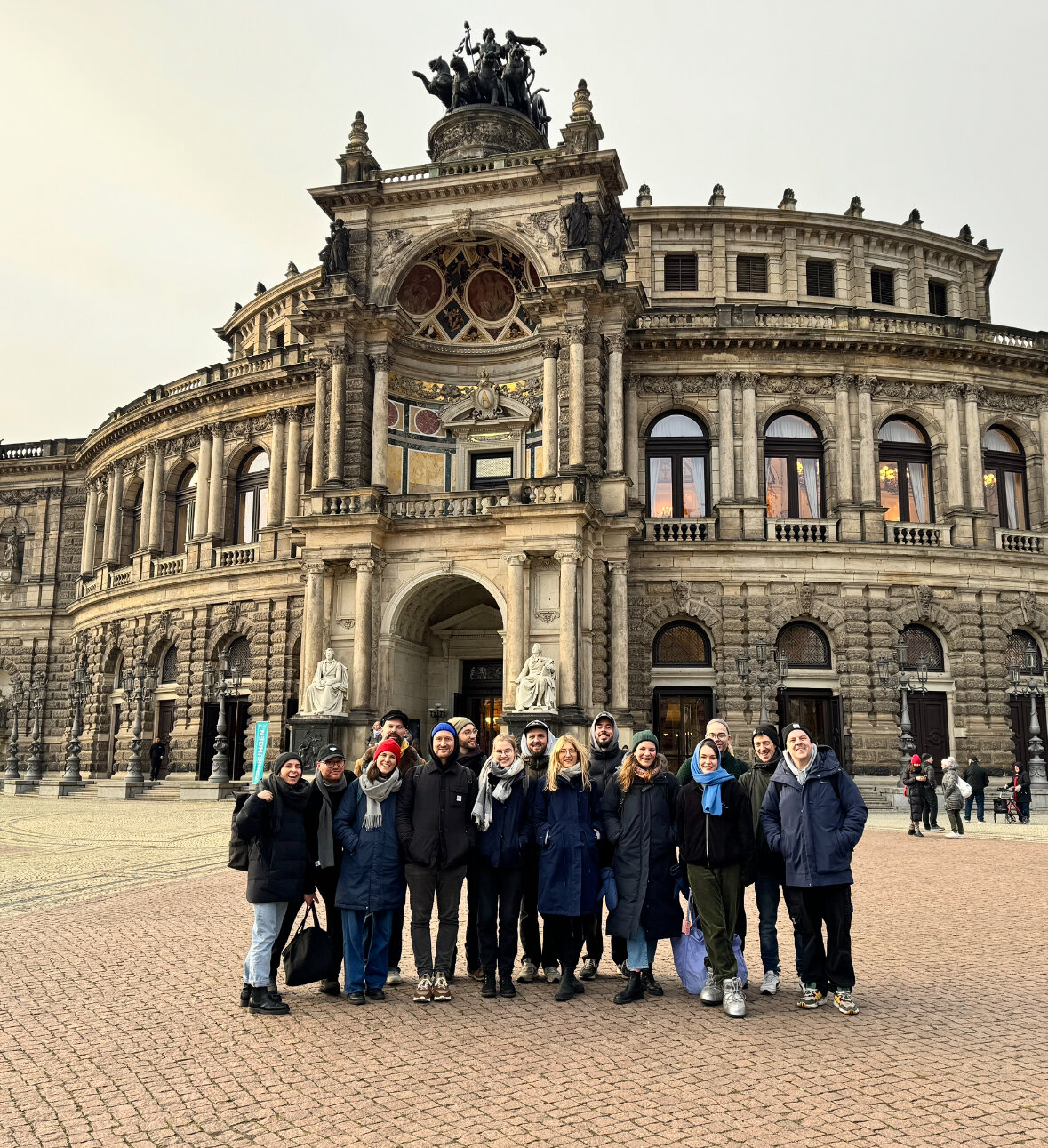Eine Gruppe von etwa zw&ouml;lf Mitarbeitenden von Studio Good steht in warmen Winterjacken vor dem historischen Semperoper-Geb&auml;ude in Dresden, einem beeindruckenden barocken Opernhaus mit gro&szlig;er Kuppel und verzierter Fassade.
