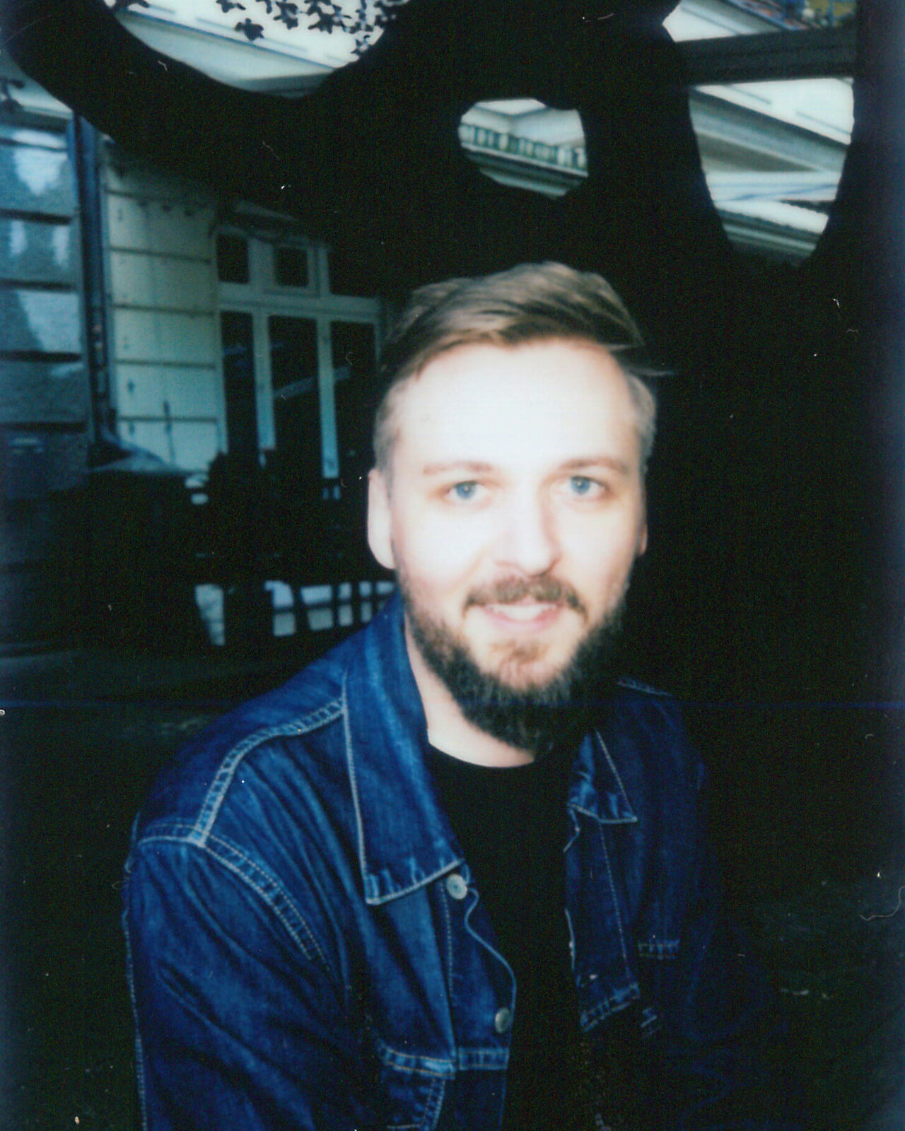 Ein Mann mit Bart in einer Jeansjacke l&auml;chelt in die Kamera, im Hintergrund ein unscharfes Geb&auml;ude.