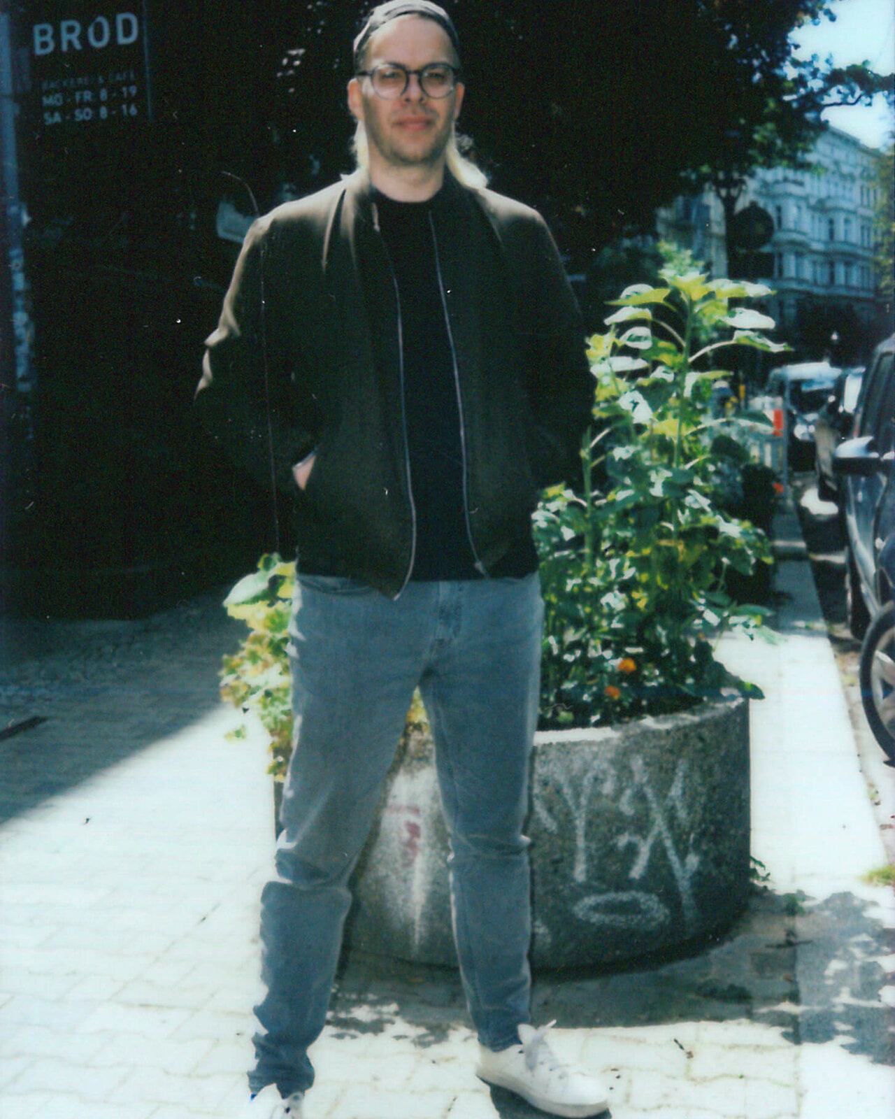 Ein Mann mit Brille und schwarzer Jacke steht auf einem Gehweg, gekleidet in blaue Jeans und wei&szlig;e Sneakers. Er posiert neben einem Blumentopf vor einem urbanen Hintergrund mit einem Schild im Hintergrund.