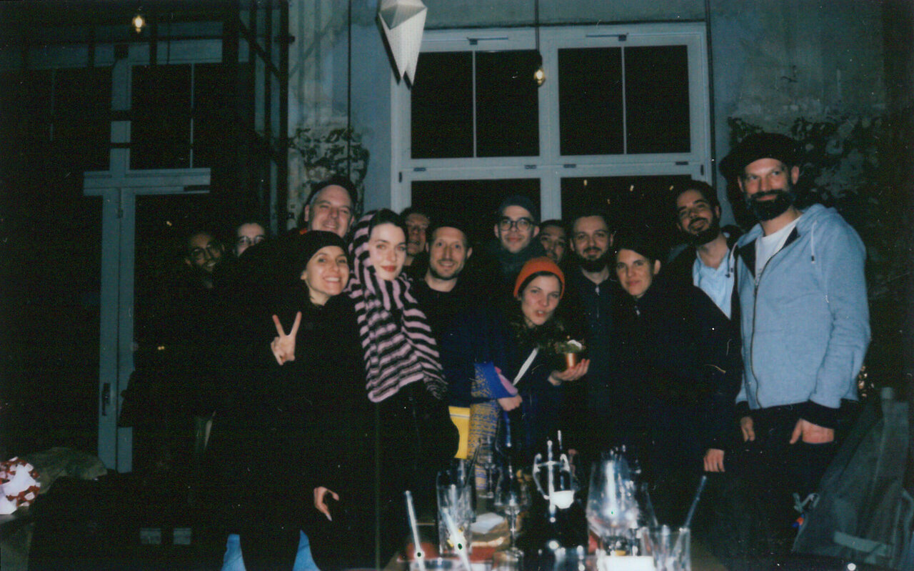 Abendliches Gruppenfoto von etwa zehn Personen bei einem Fest oder Treffen, mit Getr&auml;nken im Vordergrund und einer lockeren, freundschaftlichen Atmosph&auml;re vor einem Fenster oder Glasfront.