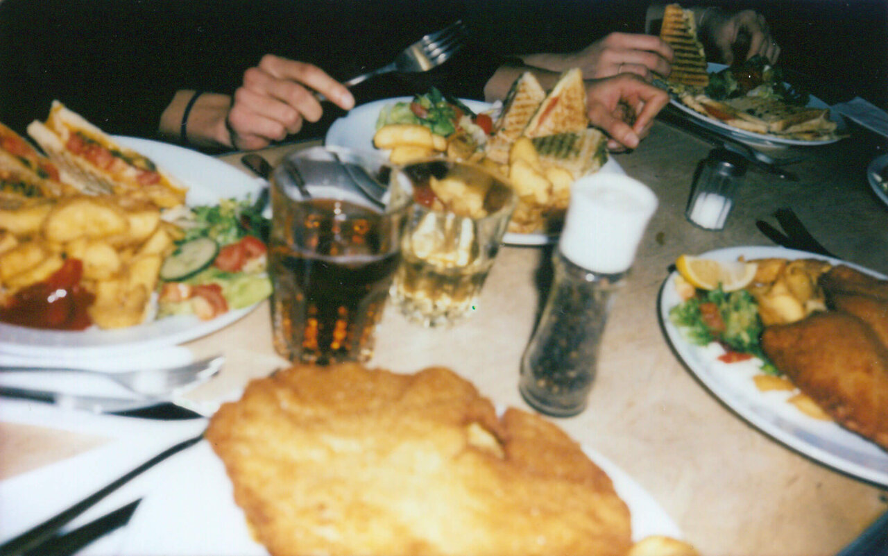 Tisch mit Tellern voller Fish and Chips, Pommes und anderen Speisen. H&auml;nde greifen nach Besteck und Gl&auml;sern mit Getr&auml;nken. Eine gesellige Mahlzeit mit Teamkollegen in einem Restaurant.