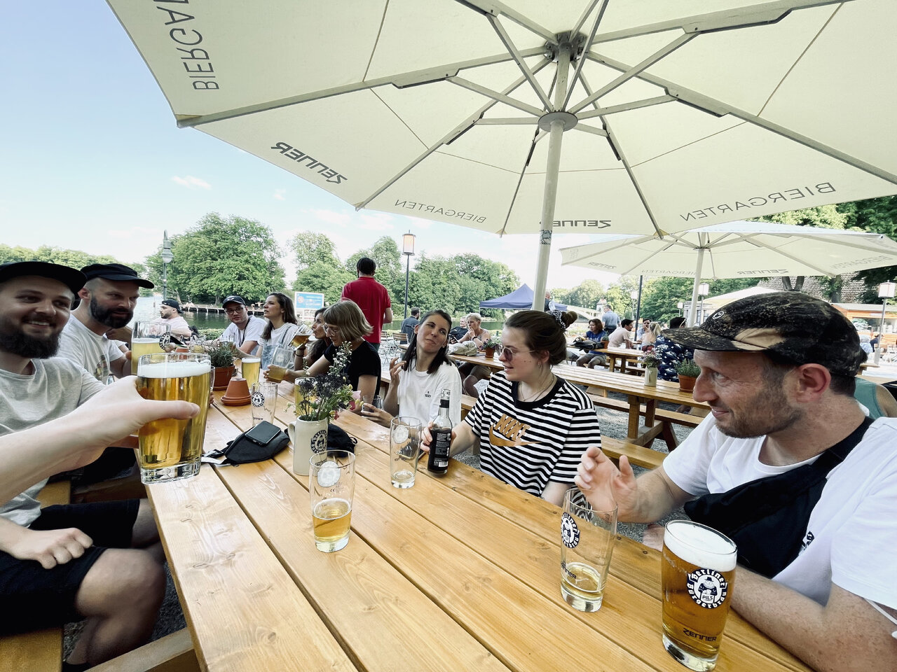 Eine Gruppe von Menschen sitzt an Holztischen unter einem gro&szlig;en Sonnenschirm und genie&szlig;t Bier und Getr&auml;nke in entspannter Atmosph&auml;re an einem sonnigen Tag.