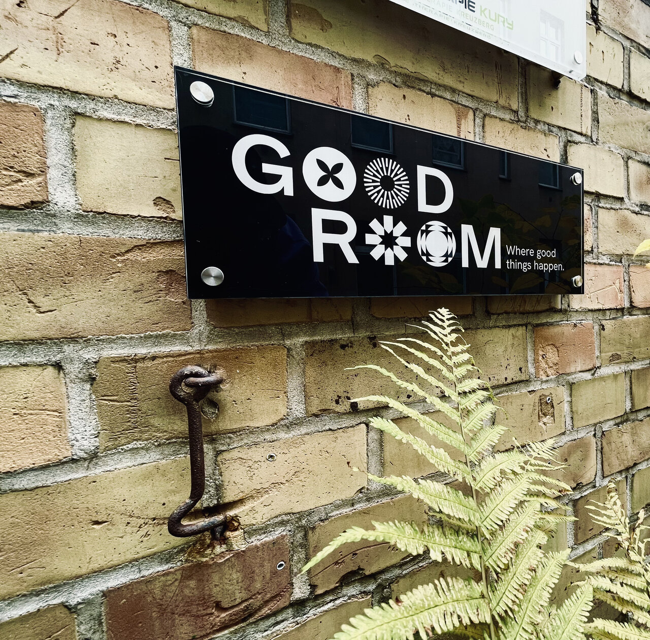 Schwarzes Schild mit "GOOD ROOM" Logo an einer beigen Ziegelwand, mit einer gr&uuml;nen Farnpflanze im Vordergrund und einem rostigen Metallhaken an der Wand.
