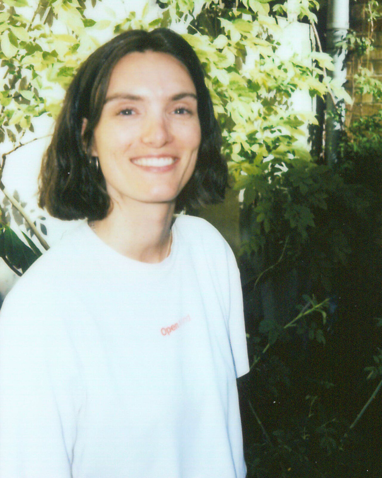 Eine Mitarbeiterin von Studio Good l&auml;chelt in die Kamera und tr&auml;gt ein wei&szlig;es T-Shirt. Sie steht vor einer gr&uuml;nen Kulisse mit Bl&auml;ttern und Zweigen, was einen nat&uuml;rlichen, entspannten Hintergrund bildet.
